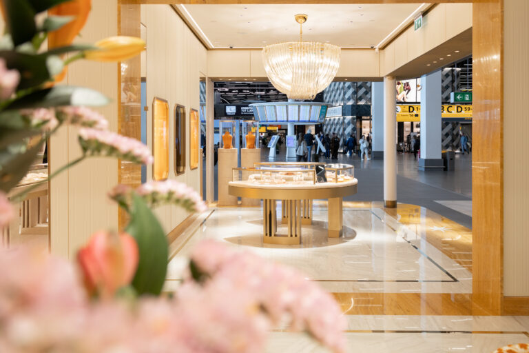 The image shows the marble interior of Schiphol Airport’s Bulgari store in Lounge 2, with products on show and pink flowers in the foreground.