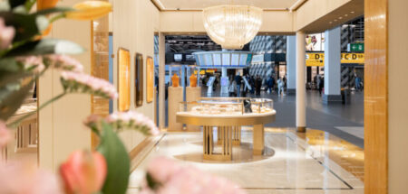 The image shows the marble interior of Schiphol Airport’s Bulgari store in Lounge 2, with products on show and pink flowers in the foreground.