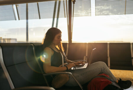 Side view of a young woman using laptop at the airport. She is working remotely while waiting to board. The light of dawn illuminates the airport.