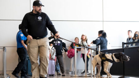A man and a dog, both in DHS uniform, patrol an airport terminal.