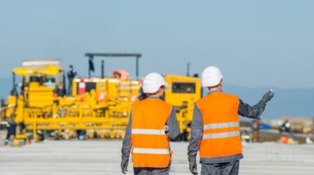 Two engineers in high-vis jackets walk down an airport runway, away from the camera gesturing to the right in conversation.
