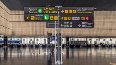 An information board inside Josep Tarradellas Barcelona-El Prat Airport stands in front of a background of ticket offices.