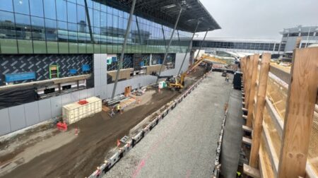 A construction site shows a wooden fence to the right and an airport terminal to the left, with a digger and some workers in high-visibility jackets working in the center.