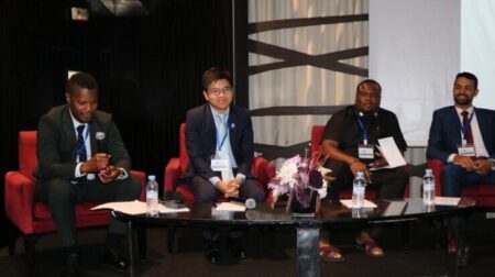 Four men in suits sit around a coffee table, which has microphones resting on it.