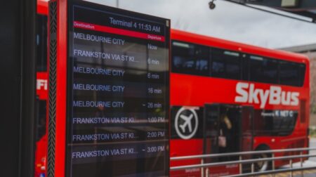 Melbourne Airport installs real-time information displays for bus services A red bus sits parked behind a display board, which shows bus times.