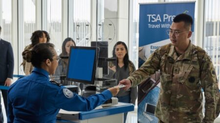 A seated TSA officer hands an ID card to a soldier in camo uniform.