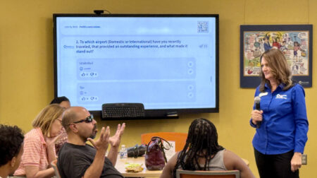 A Denver INternational Airport staff member stands on a stage with a digital screen in front of seated audience members