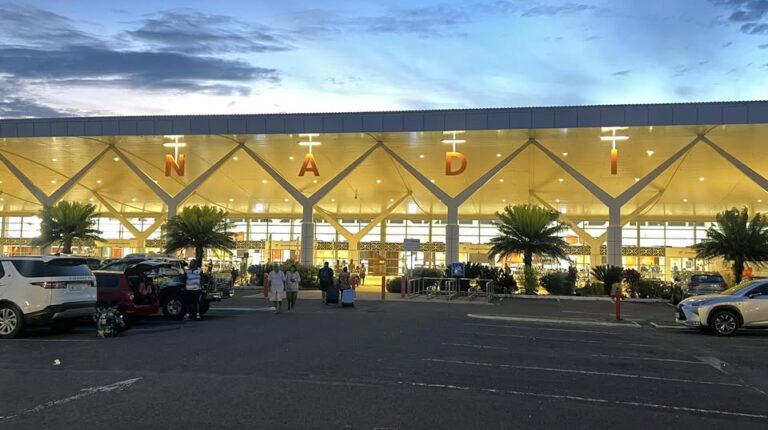 The entrance to Nadi International Airport in Fiji at dusk, with the frontage illuminated by bright yellow lighting and drakening skies above