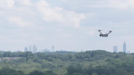 A Flock drone flies over an infrastructure site with green trees and buildings in the foreground and skyscrapers along the horizon