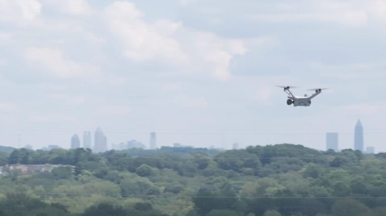 A Flock drone flies over an infrastructure site with green trees and buildings in the foreground and skyscrapers along the horizon