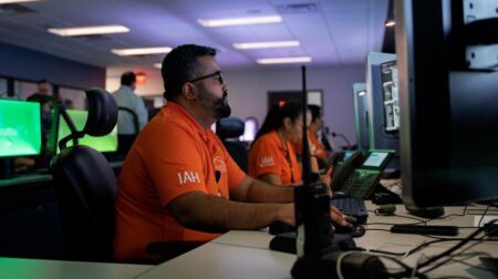 Security staff sit in front of a screen at a large desk of multiple workstations in a busy command center room
