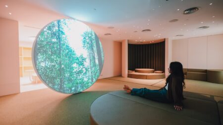 The interior of the sensory-friendly Calm Room at Changi Airport with soft lighting and surfaces and a large circular light depicting a forest scene in the middle of the room. A traveler sits on the floor with their legs stretched out flat in front of them.