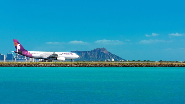 A Hawaiian Airlines jet taxis down the runway at a Hawai‘i airport, with a blue sea in the foreground and a blue sky above.