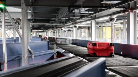 Houston Hobby and Melbourne Orlando to upgrade BHS Baggage handling system in an airport, with luggage being transported on conveyor belts. There is a red, a silver and other darker coloured bags in the background.