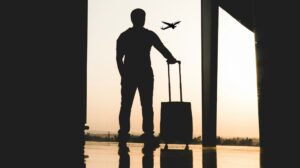A man with a small piece of luggage on wheels stands in an airport terminal silhouetted against the sky as a plane takes off in the background.