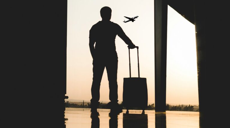 A man with a small piece of luggage on wheels stands in an airport terminal silhouetted against the sky as a plane takes off in the background.