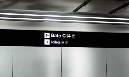 New signing by Astra Group at Manchester Airport Terminal 2. The dark sign is on a silver wall and says 'Gate C14' with an arrow pointing to the left, and 'Toilets' with an arrow pointing to the right.