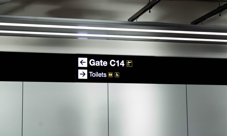 New signing by Astra Group at Manchester Airport Terminal 2. The dark sign is on a silver wall and says 'Gate C14' with an arrow pointing to the left, and 'Toilets' with an arrow pointing to the right.