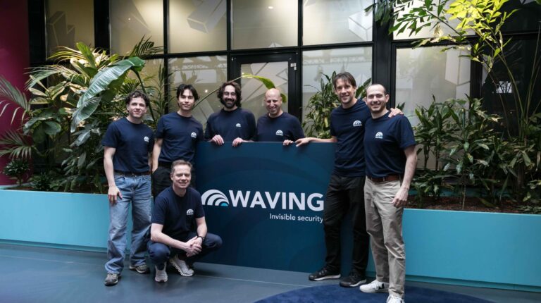Waving staff in dark blue company-branded clothing stand in front of a branded sign at Schiphol airport in Amsterdam.