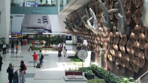 the interior or a large airport with green foliage, a large art installation and passengers walking across the concourse.