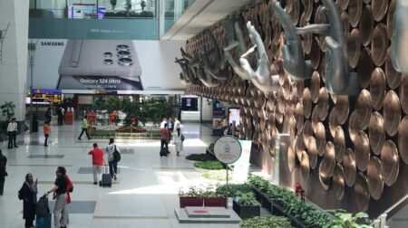 the interior or a large airport with green foliage, a large art installation and passengers walking across the concourse.