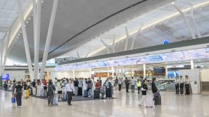Departures terminal concourse at Hong Kong Airport's new Terminal 2. Passengers are queueing in lines for check-in and bag drop.