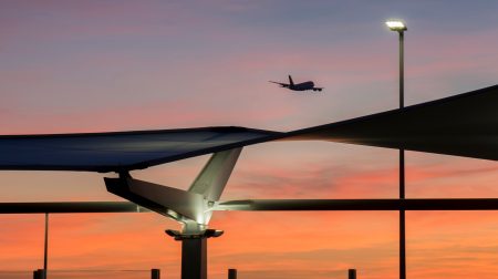 A plane takes off at an airport at night time as the sun sets. Airport infrastructure is visible in silhouette in the foreground.