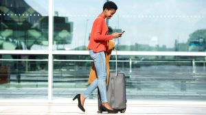 A woman wearing a red jacket and light blue trousers walks through an airport pulling a small piece of luggage.
