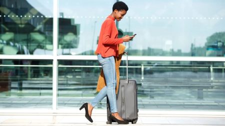 A woman wearing a red jacket and light blue trousers walks through an airport pulling a small piece of luggage.