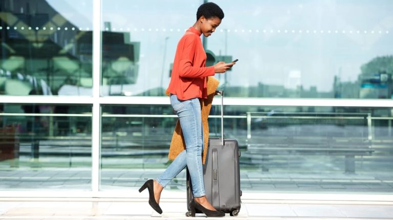 A woman wearing a red jacket and light blue trousers walks through an airport pulling a small piece of luggage.