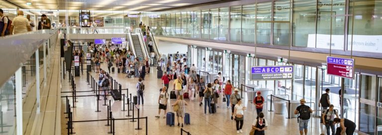 Concourse at Geneve Airport, with passengers moving to their gates. There is an escalator and stairs to an upper level.