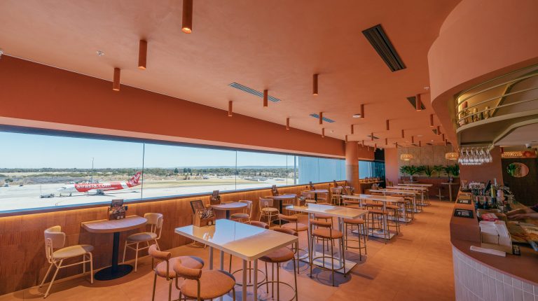 Perth Airport unveils refreshed T1 International retail precinct ahead of winter travel surge Co + Co at Perth Airport T1 International terminal. It has a wooden tables and chairs, a long window with views of the runways, and hanging lights.