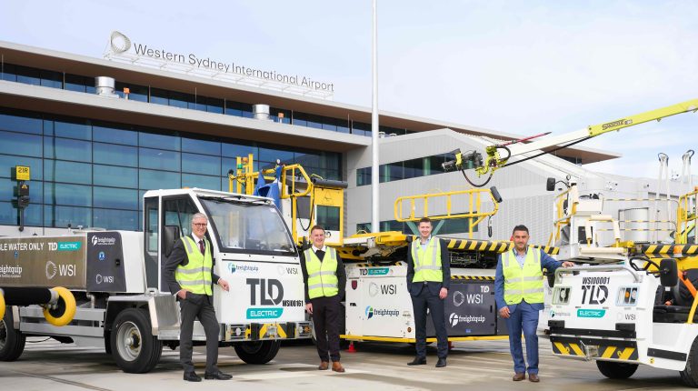 Representatives of Western Sydney Airport stand on the tarmac among ground operations equipment, wearing yellow high-vis jackets.