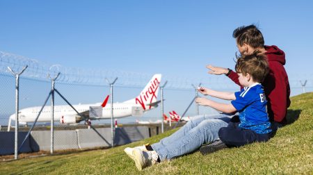Brisbane Airport opens plane-spotting area, Fellowship Place Two people sit on the grass at Brisbane Airport's new plane-spotting area, Fellowship Place, watching a Virgin airplane on the tarmac.