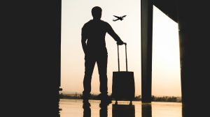 A man with a small piece of rolling luggage stands silhouetted against a window looking out of the tarmac at an airport as the sun sets.