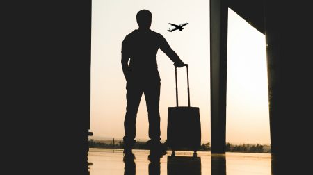 A man with a small piece of rolling luggage stands silhouetted against a window looking out of the tarmac at an airport as the sun sets.