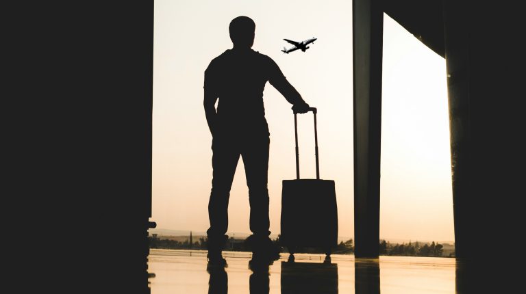 A man with a small piece of rolling luggage stands silhouetted against a window looking out of the tarmac at an airport as the sun sets.