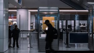 A security checkpoint at an airport. A man walks past in silhouette in the foreground.
