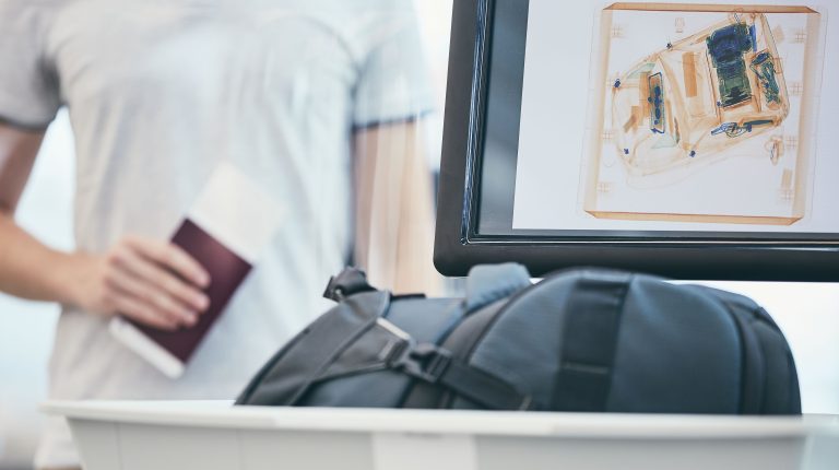 A person wearing a white t-shirt holds their passport in their right hand as they put a dark bag into a security screening tray.