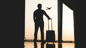 Silhouette of a man standing with a small piece of rolling luggage by a window in an airport terminal as a plane flies in the distance.