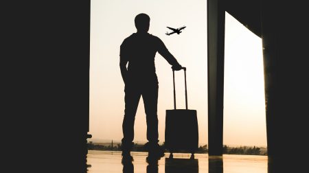 Silhouette of a man standing with a small piece of rolling luggage by a window in an airport terminal as a plane flies in the distance.