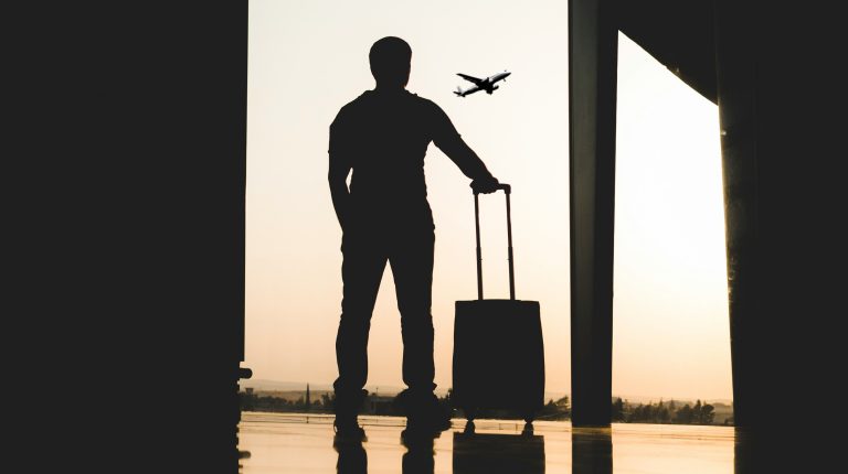 Silhouette of a man standing with a small piece of rolling luggage by a window in an airport terminal as a plane flies in the distance.
