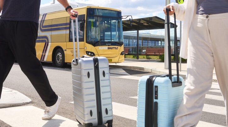 Yellow shuttle bus at bus stop with two passengers wheeling suitcases in foreground.